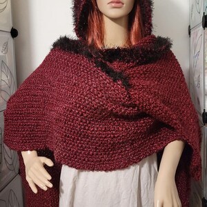 Pu&ograve; includere: Un mantello con cappuccio bordeaux con finiture in ecopelliccia nera. Il mantello &egrave; realizzato in un tessuto a maglia strutturato e cade sulle spalle. Anche il cappuccio &egrave; rifinito con ecopelliccia nera. Il mantello &egrave; esposto su un manichino.