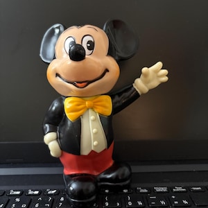 May include: A vintage black and yellow Mickey Mouse figurine with a red and white tuxedo. The figurine is standing on a black laptop keyboard.