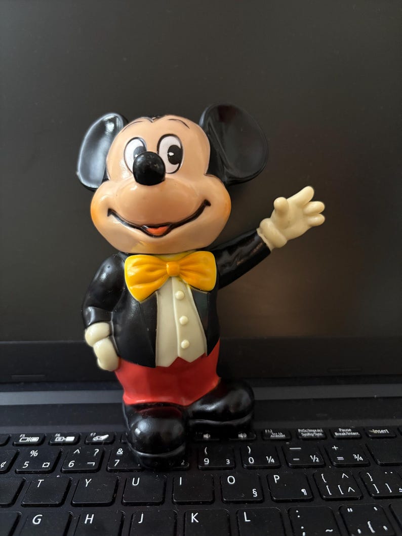May include: A vintage black and yellow Mickey Mouse figurine with a red and white tuxedo. The figurine is standing on a black laptop keyboard.