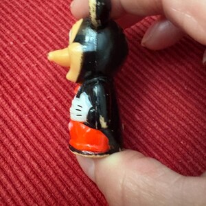 May include: A small, vintage Mickey Mouse figurine. The figure has a black body, a yellow beak, and red shorts. The figure is held against a red background.
