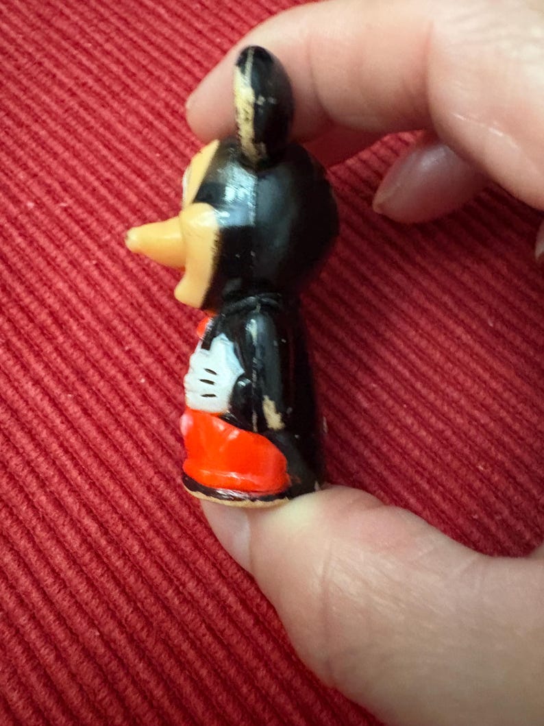 May include: A small, vintage Mickey Mouse figurine. The figure has a black body, a yellow beak, and red shorts. The figure is held against a red background.