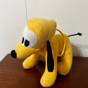 Pu&ograve; includere: Un cane di peluche giallo con orecchie nere e naso nero. Il cane ha occhi bianchi e un collare giallo.