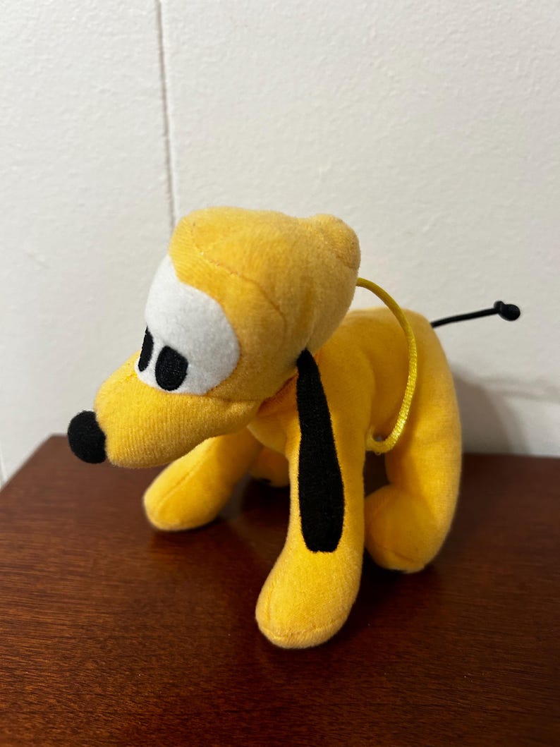 Pu&ograve; includere: Un cane di peluche giallo con orecchie nere e naso nero. Il cane ha occhi bianchi e un collare giallo.