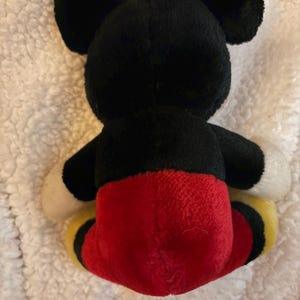 May include: A plush toy of a famous cartoon mouse, viewed from the back. The toy has black ears, head, and arms, with a red bottom and yellow and black accents on the feet. The toy is on a white, textured surface.