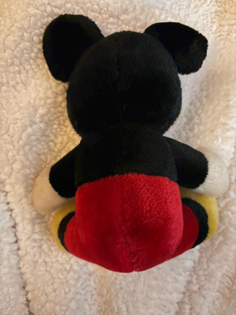 May include: A plush toy of a famous cartoon mouse, viewed from the back. The toy has black ears, head, and arms, with a red bottom and yellow and black accents on the feet. The toy is on a white, textured surface.