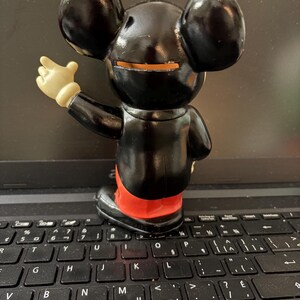 May include: A black plastic Mickey Mouse figurine with a red and white outfit. The figurine is standing on a black laptop keyboard.