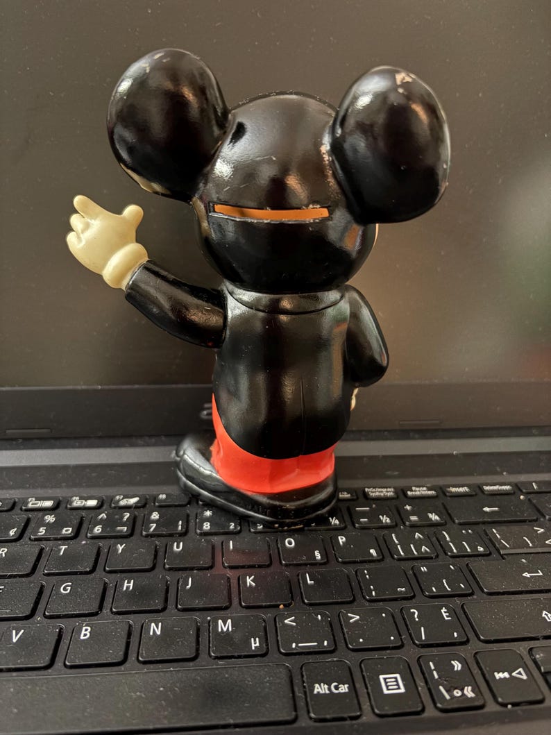 May include: A black plastic Mickey Mouse figurine with a red and white outfit. The figurine is standing on a black laptop keyboard.