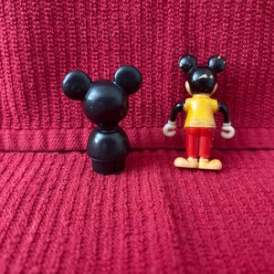 May include: Two black plastic Mickey Mouse figurines. One is a simple, standing figure. The other is a more detailed figure with red pants and a yellow shirt.