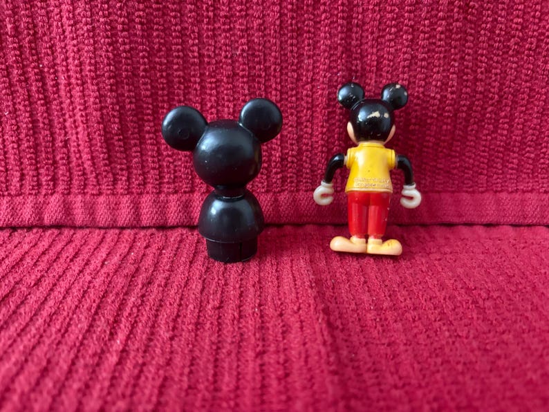 May include: Two black plastic Mickey Mouse figurines. One is a simple, standing figure. The other is a more detailed figure with red pants and a yellow shirt.