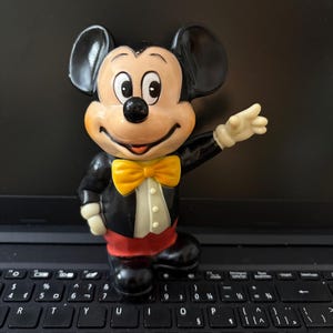 May include: A black and yellow plastic Mickey Mouse figurine with a yellow bow tie. The figurine is standing on a black laptop keyboard.