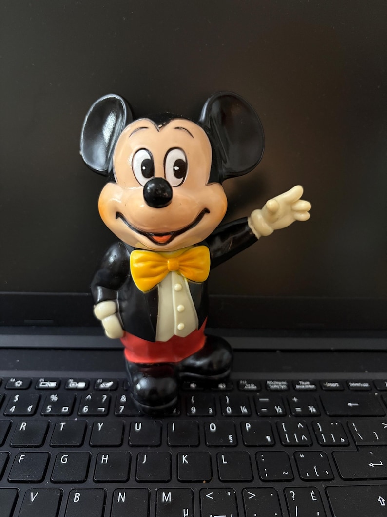 May include: A black and yellow plastic Mickey Mouse figurine with a yellow bow tie. The figurine is standing on a black laptop keyboard.
