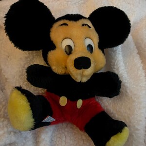 May include: A plush Mickey Mouse toy with black ears, arms, and legs, a yellow face, and red shorts with yellow buttons. The toy is lying on a white, textured surface.