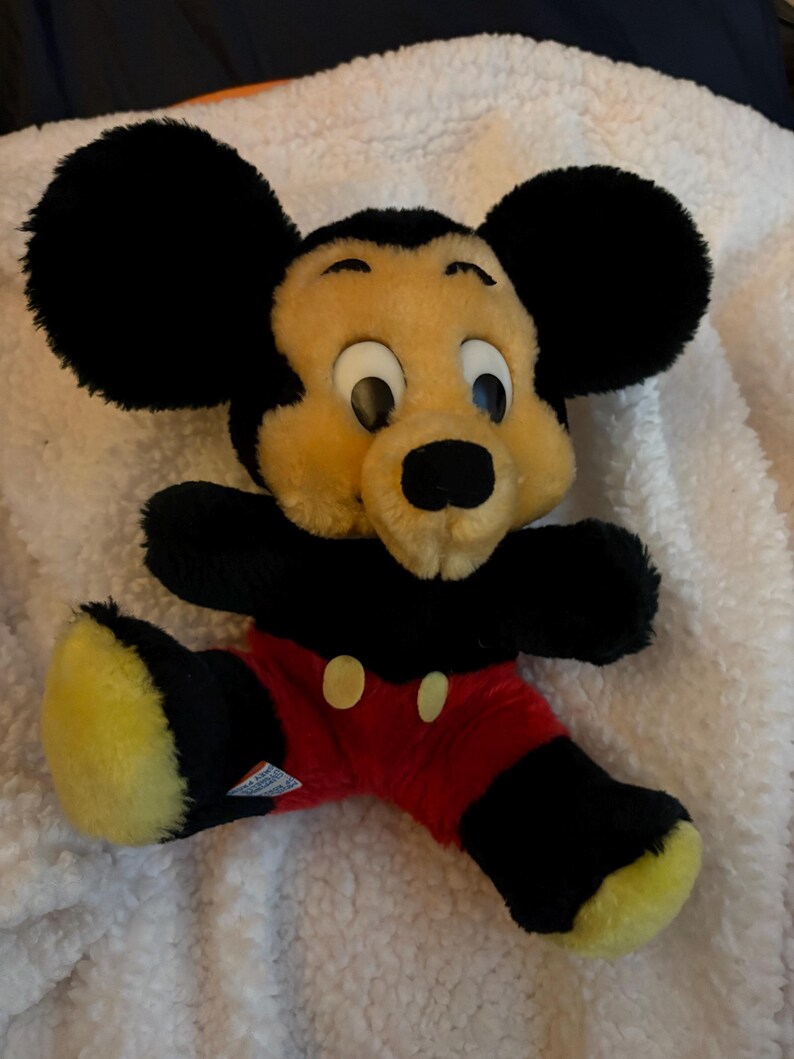 May include: A plush Mickey Mouse toy with black ears, arms, and legs, a yellow face, and red shorts with yellow buttons. The toy is lying on a white, textured surface.