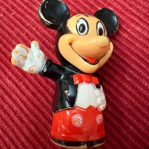 May include: Vintage Mickey Mouse figurine. The figure is wearing a black tuxedo jacket, red shorts, and a red bow tie. The figure has white gloves and a smiling expression. The figurine is on a red background.