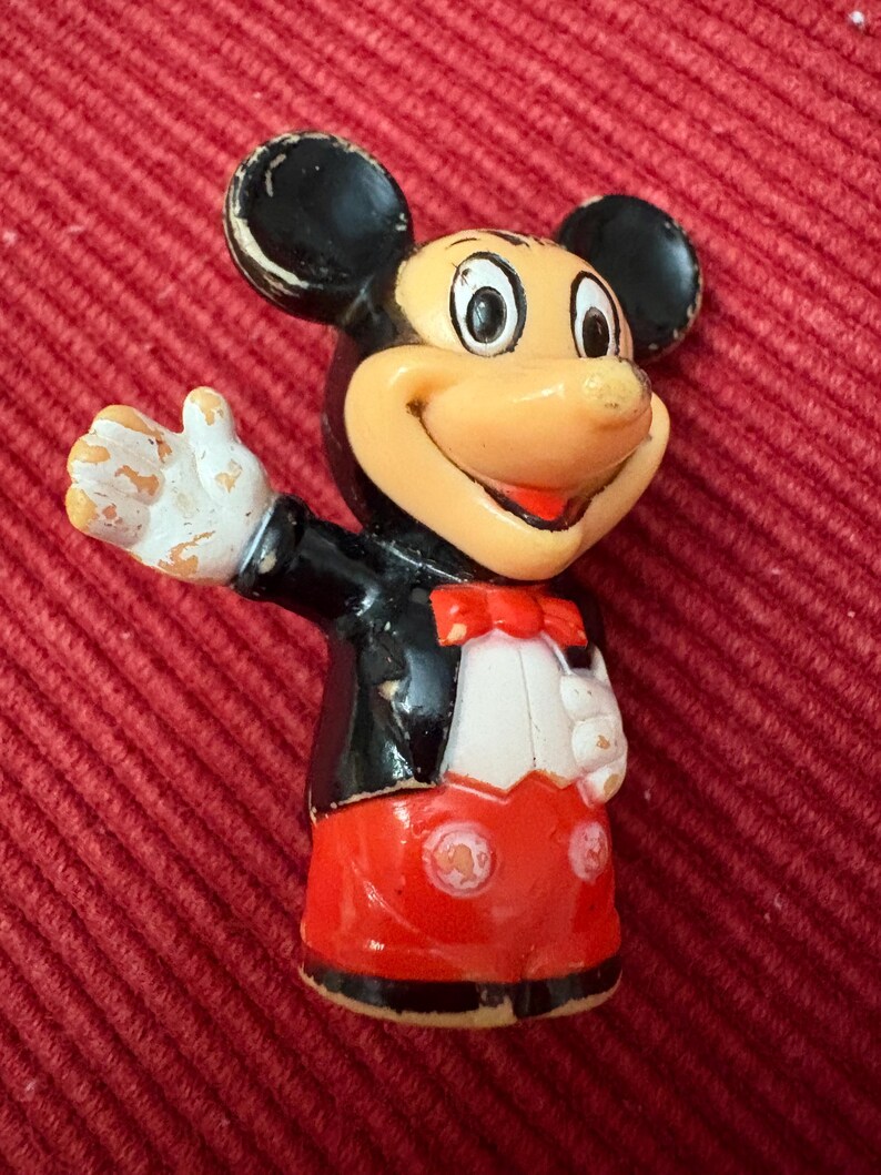 May include: Vintage Mickey Mouse figurine. The figure is wearing a black tuxedo jacket, red shorts, and a red bow tie. The figure has white gloves and a smiling expression. The figurine is on a red background.