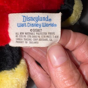 May include: A close-up of a plush toy tag with the text "Disneyland Walt Disney World" and "DISNEY". The tag also includes details about materials and manufacturing, including "PRODUCT OF SRILANKA". The toy is red, black, and yellow.