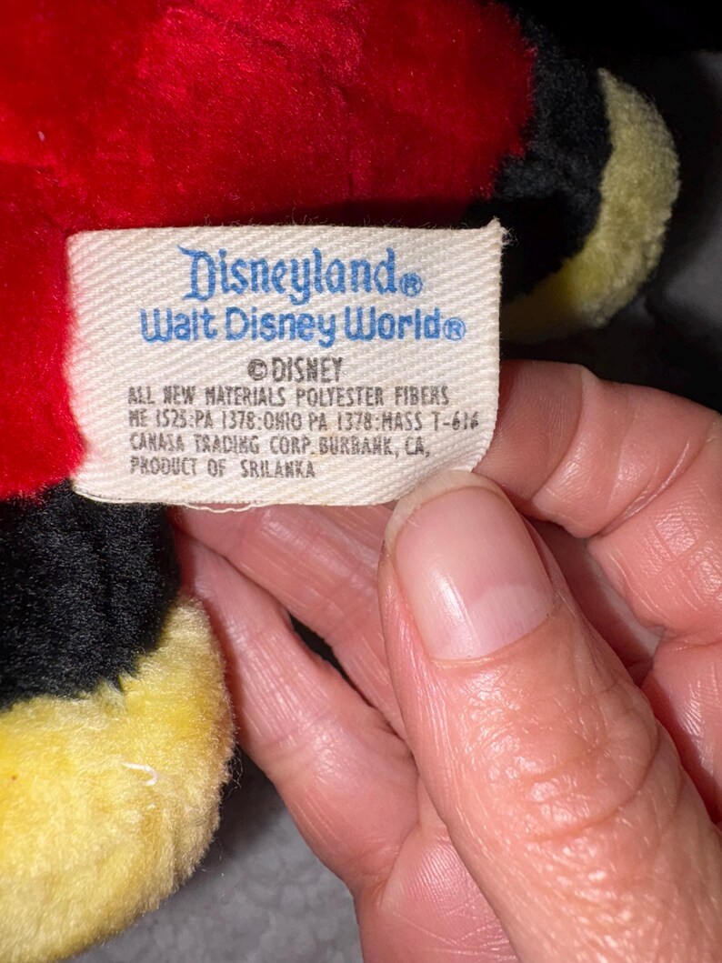 May include: A close-up of a plush toy tag with the text "Disneyland Walt Disney World" and "DISNEY". The tag also includes details about materials and manufacturing, including "PRODUCT OF SRILANKA". The toy is red, black, and yellow.