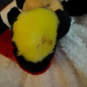 May include: Close-up of a plush toy featuring a yellow beak, black ears, and a red and black hat. The toy is set against a white, textured background. The toy's design is reminiscent of a cartoon character.