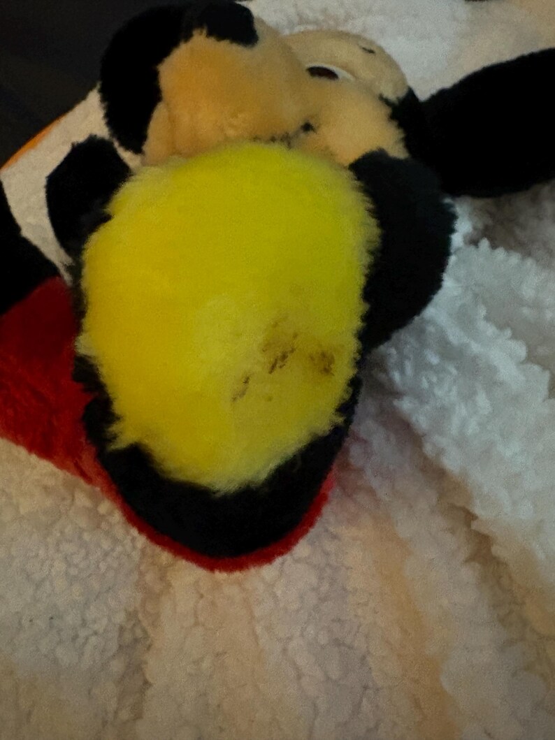 May include: Close-up of a plush toy featuring a yellow beak, black ears, and a red and black hat. The toy is set against a white, textured background. The toy's design is reminiscent of a cartoon character.