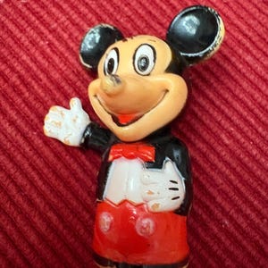 May include: A vintage Mickey Mouse figurine. The figure has a tan face, black ears, and a red bow tie. Mickey is wearing a black jacket, white gloves, and red shorts. The figurine is posed with one arm raised in a friendly gesture.