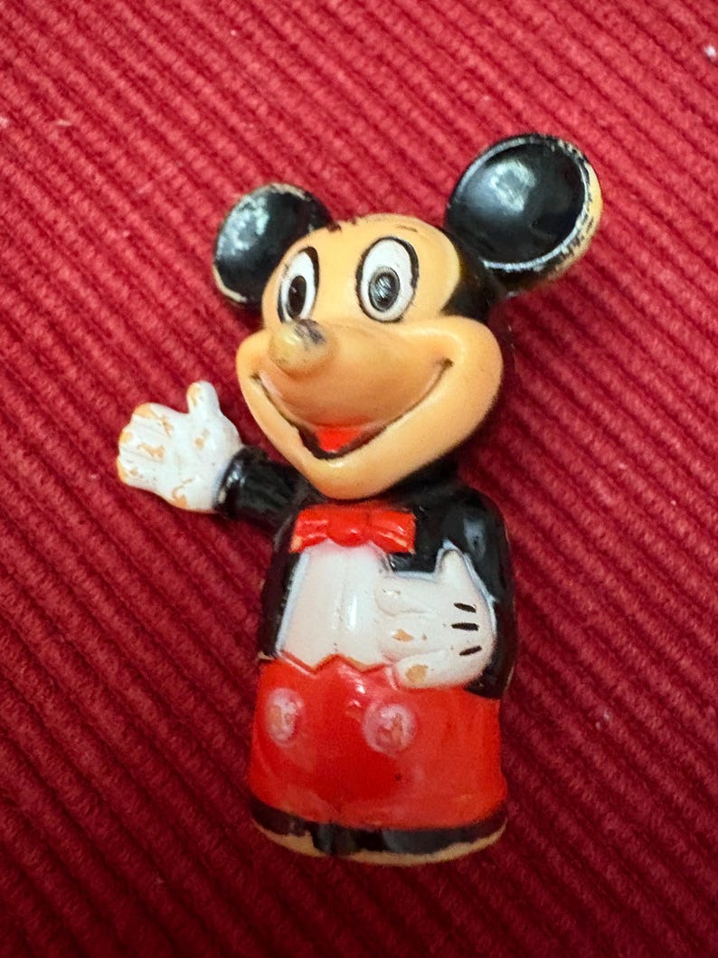 May include: A vintage Mickey Mouse figurine. The figure has a tan face, black ears, and a red bow tie. Mickey is wearing a black jacket, white gloves, and red shorts. The figurine is posed with one arm raised in a friendly gesture.