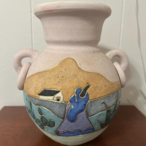 Peut inclure: Un vase en céramique rose avec deux anses, décoré d'une scène peinte d'une maison, d'une figure et de cactus. Le vase a une surface texturée et un aspect rustique peint à la main.