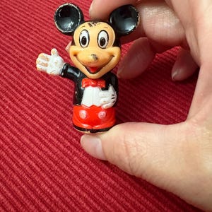 May include: A small Mickey Mouse figurine, featuring black ears, a tan face, and a red bow tie. Mickey wears a black jacket, white gloves, and red shorts. The figurine is held against a red background.