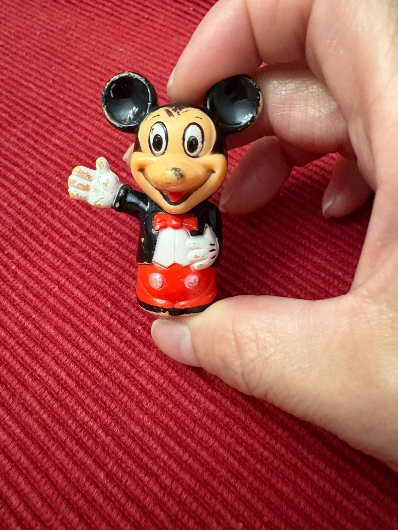 May include: A small Mickey Mouse figurine, featuring black ears, a tan face, and a red bow tie. Mickey wears a black jacket, white gloves, and red shorts. The figurine is held against a red background.