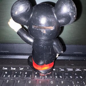 Pu&ograve; includere: Salvadanaio di plastica nero a forma di Topolino con uno slot sul retro per le monete. Il salvadanaio &egrave; appoggiato su una tastiera di laptop nera.