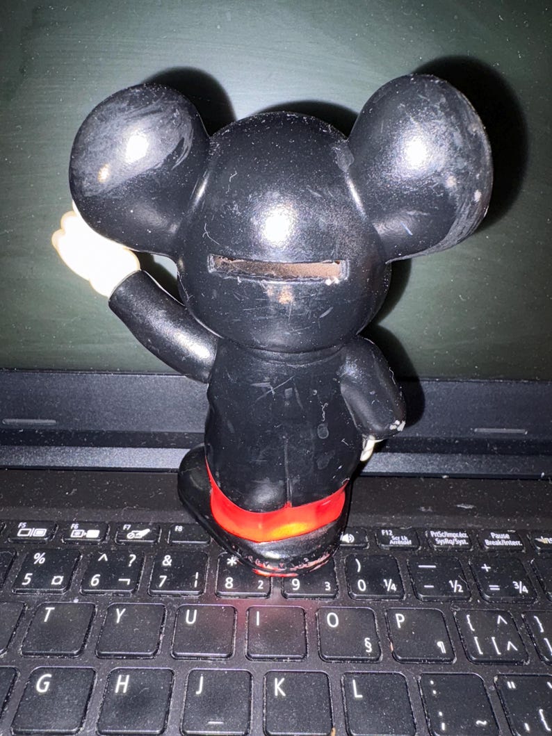 Pu&ograve; includere: Salvadanaio di plastica nero a forma di Topolino con uno slot sul retro per le monete. Il salvadanaio &egrave; appoggiato su una tastiera di laptop nera.