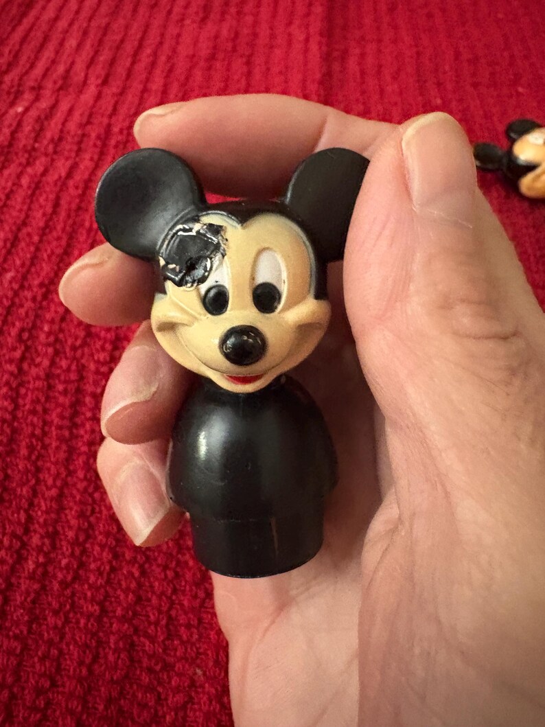 May include: A black and yellow plastic Mickey Mouse figurine with one eye missing. The figurine is wearing a black shirt and has a black nose and ears.