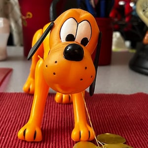 Könnte beinhalten: Ein orangefarbener Plastik-Spielzeug-Hund mit schwarzen Ohren und einer schwarzen Nase. Der Hund steht auf einer roten Oberfläche mit einem goldenen Etikett, auf dem "Pluto" steht.