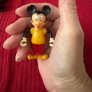 May include: A vintage plastic Mickey Mouse figurine with black ears, yellow shirt, and red pants. The figurine is holding a white circle in each hand.