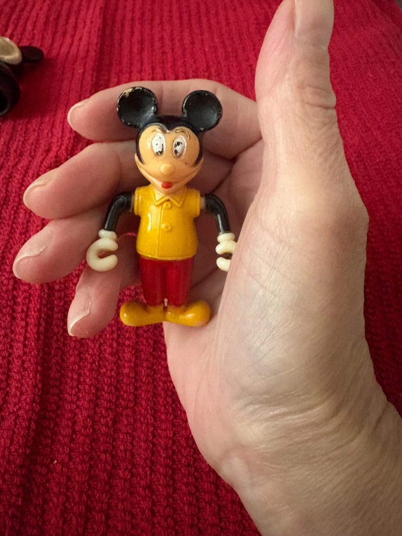 May include: A vintage plastic Mickey Mouse figurine with black ears, yellow shirt, and red pants. The figurine is holding a white circle in each hand.