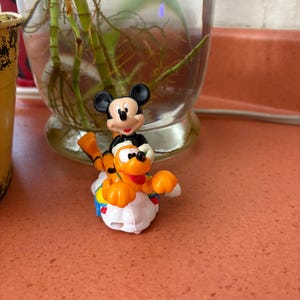 Könnte beinhalten: Eine kleine Mickey Mouse-Figur reitet auf einer Pluto-Figur. Mickey Mouse trägt ein schwarzes Outfit mit weißen Handschuhen und einem roten Lächeln. Pluto ist orange und gelb und sitzt auf einer weißen wolkenförmigen Basis.