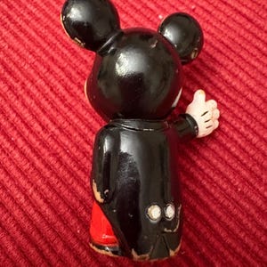 May include: A vintage Mickey Mouse figurine, viewed from the back. The figure is primarily black with red shorts and white gloves. The figurine is giving a thumbs up. The figure has two large black ears.