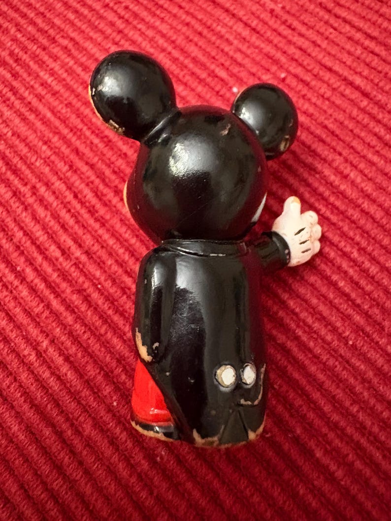 May include: A vintage Mickey Mouse figurine, viewed from the back. The figure is primarily black with red shorts and white gloves. The figurine is giving a thumbs up. The figure has two large black ears.