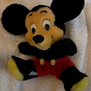 May include: A vintage Mickey Mouse plush toy. The plush features a yellow face, black ears, and black and red clothing. The toy is lying on a white, textured surface.