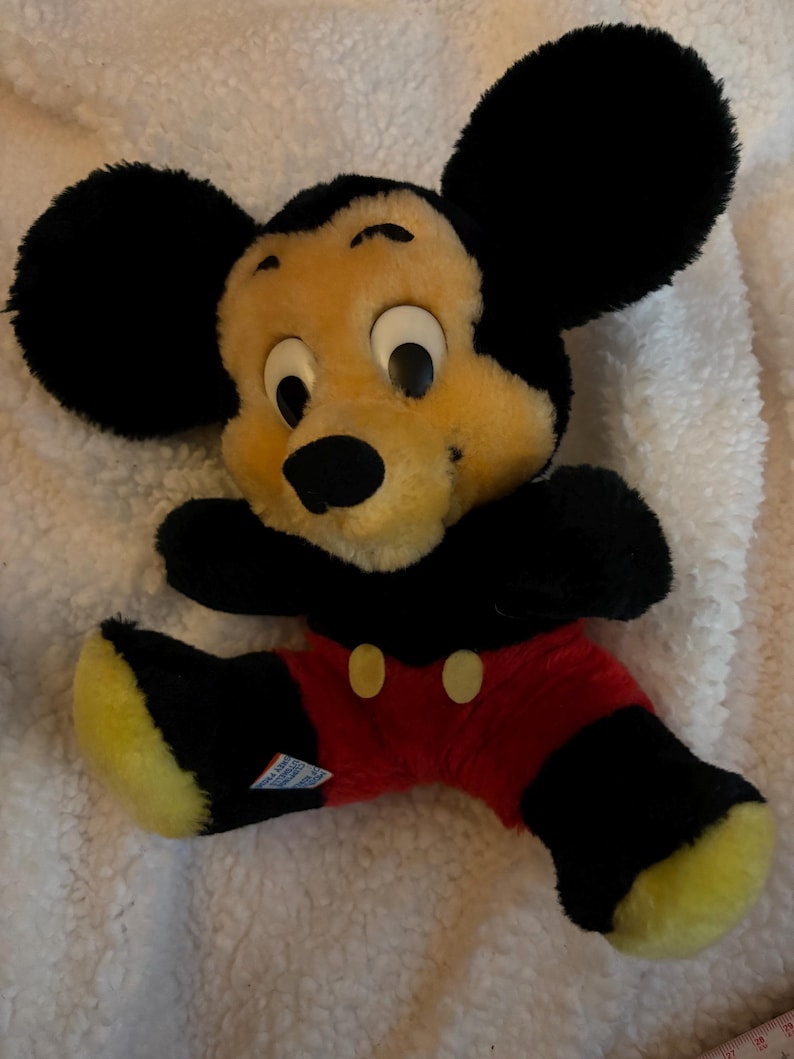 May include: A vintage Mickey Mouse plush toy. The plush features a yellow face, black ears, and black and red clothing. The toy is lying on a white, textured surface.
