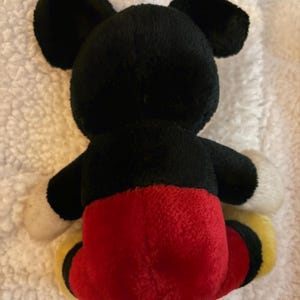May include: A plush Mickey Mouse toy, viewed from the back. The toy features black ears and head, white gloves, red shorts, and yellow shoes. The toy is made of soft, fuzzy material.