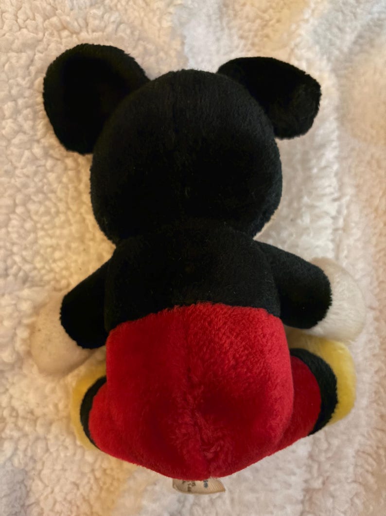 May include: A plush Mickey Mouse toy, viewed from the back. The toy features black ears and head, white gloves, red shorts, and yellow shoes. The toy is made of soft, fuzzy material.