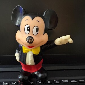 Pu&ograve; includere: Una statuetta vintage di Topolino in plastica che indossa uno smoking nero con un papillon giallo. La statuetta ha una base rossa e nera e si trova su una tastiera di un laptop nero.