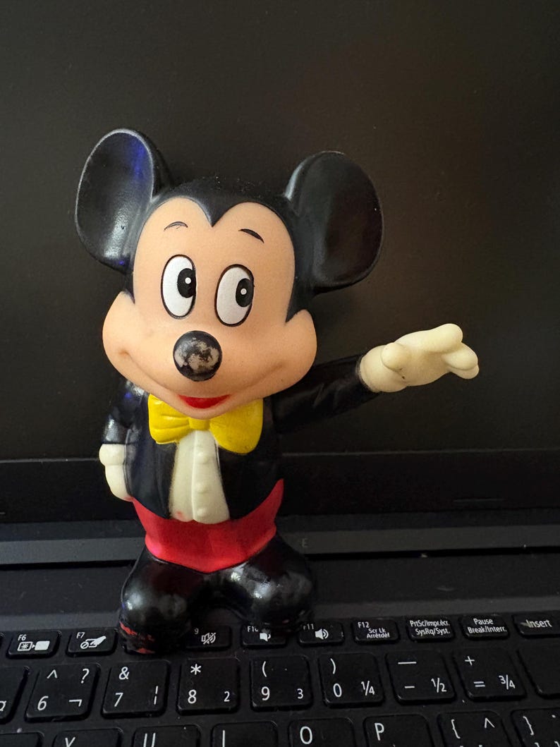 Pu&ograve; includere: Una statuetta vintage di Topolino in plastica che indossa uno smoking nero con un papillon giallo. La statuetta ha una base rossa e nera e si trova su una tastiera di un laptop nero.