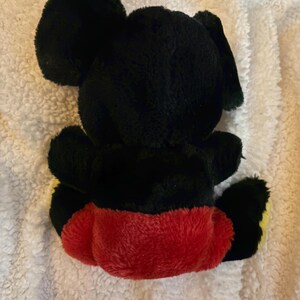 May include: A plush toy in the shape of a cartoon mouse, viewed from the back. The toy is primarily black with large, rounded ears. It has a red bottom and yellow accents on the feet. The toy is sitting on a white, textured surface.