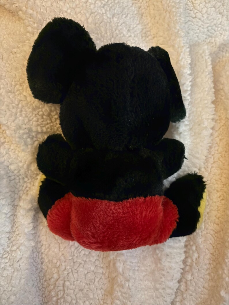 May include: A plush toy in the shape of a cartoon mouse, viewed from the back. The toy is primarily black with large, rounded ears. It has a red bottom and yellow accents on the feet. The toy is sitting on a white, textured surface.