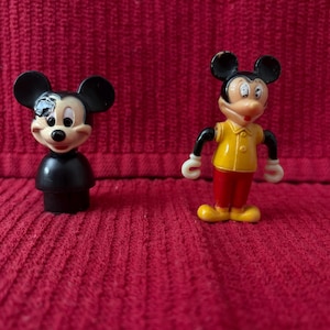 May include: Two plastic Mickey Mouse figurines. One is black with a red nose and the other is yellow, red, and black with a white shirt.