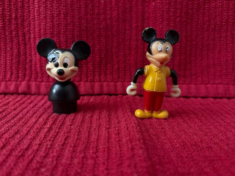 May include: Two plastic Mickey Mouse figurines. One is black with a red nose and the other is yellow, red, and black with a white shirt.