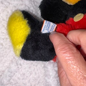 May include: A vintage Mickey Mouse plush toy, featuring black fur, a yellow face and feet, and a red body. The toy has a tag that reads "MOUSE OF KOREA CLIPPINGS IT SHELLS HEY PRODS."