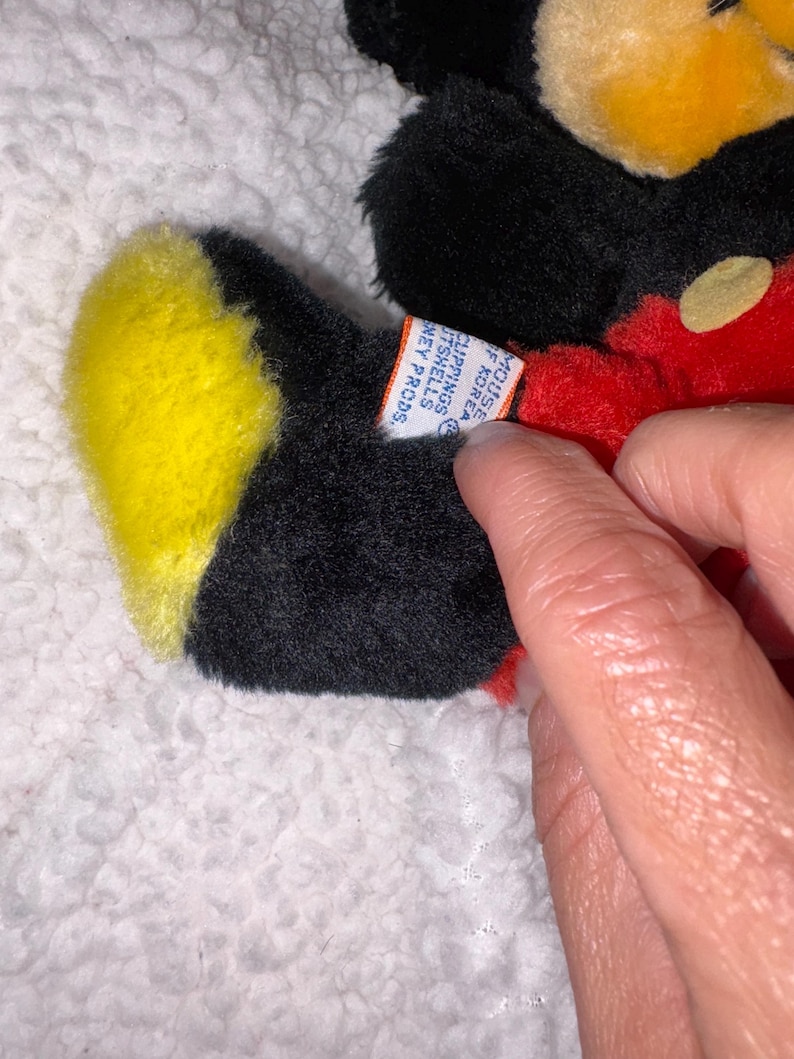 May include: A vintage Mickey Mouse plush toy, featuring black fur, a yellow face and feet, and a red body. The toy has a tag that reads "MOUSE OF KOREA CLIPPINGS IT SHELLS HEY PRODS."