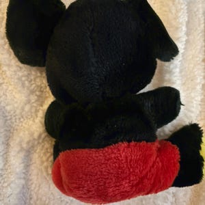 May include: A plush toy of a cartoon mouse, viewed from the back. The toy has black fur, large round ears, and red shorts. The toy's feet are yellow. The toy is sitting on a white, textured surface.